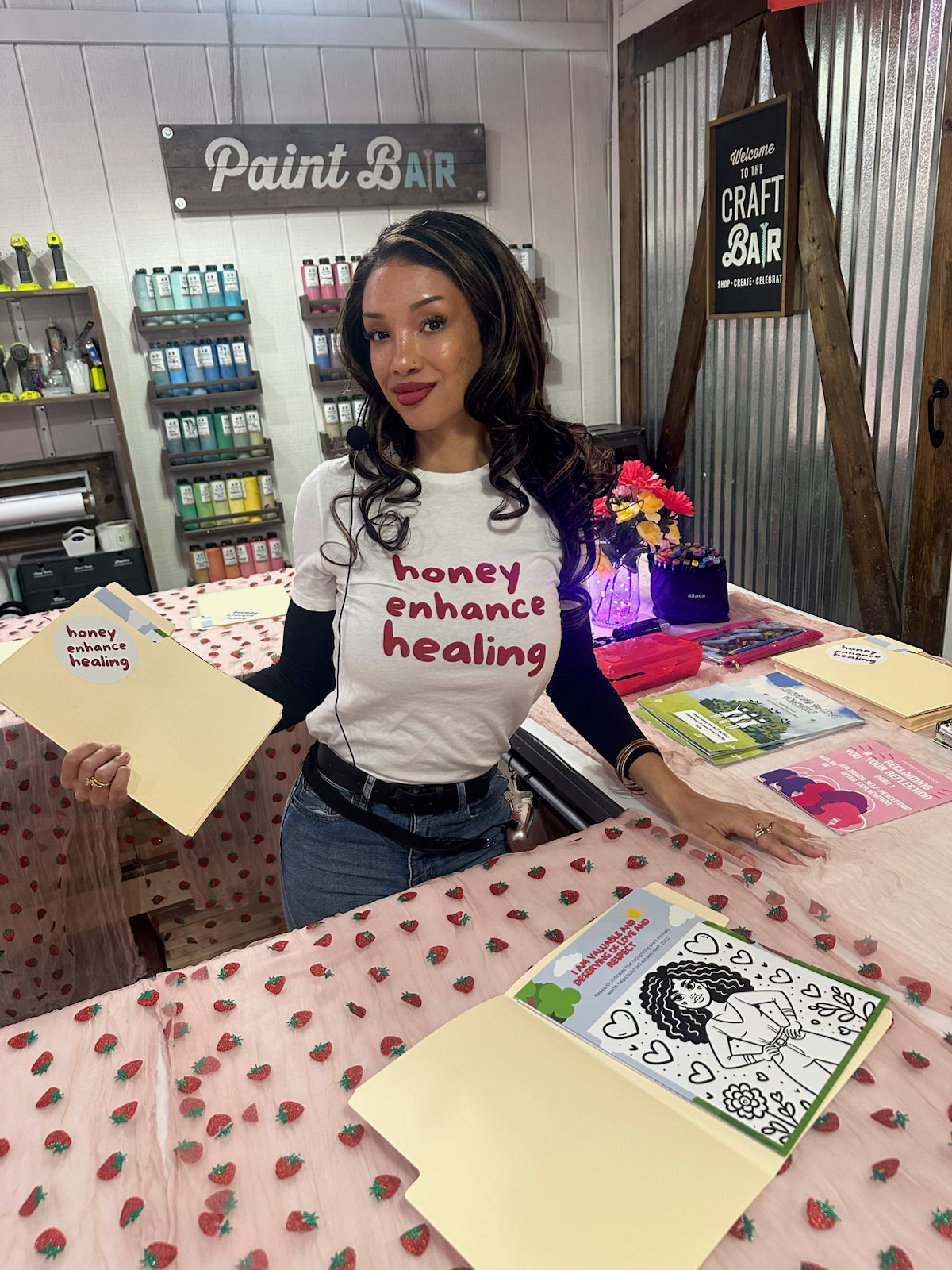 Woman Founder in a white Honey Enhance Healing t-shirt at a craft fair table with healing coloring book sheets and journaling supplies.